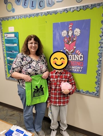 UGI employee holds Ready.Set.Read bag and stands next to the student she tutors. Student's face is covered with a smiley face emoji due to photo permissions.