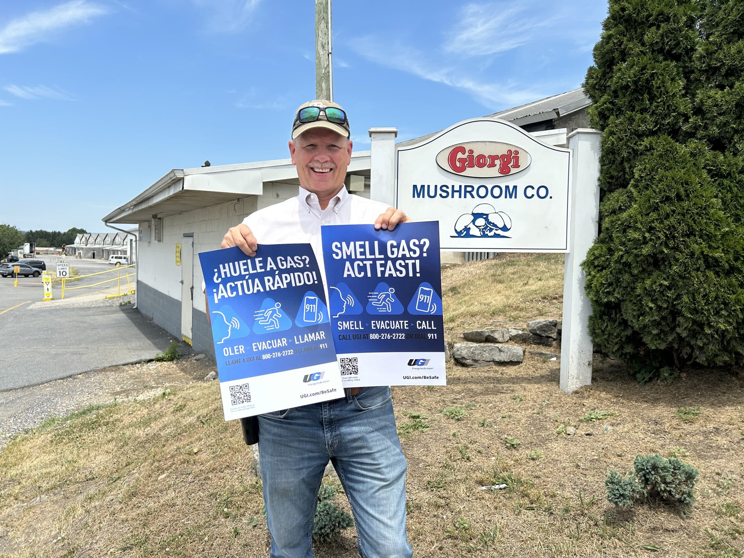 Man holds up English and Spanish posters with gas safety information in front of Giorgi Mushroom Co. sign.