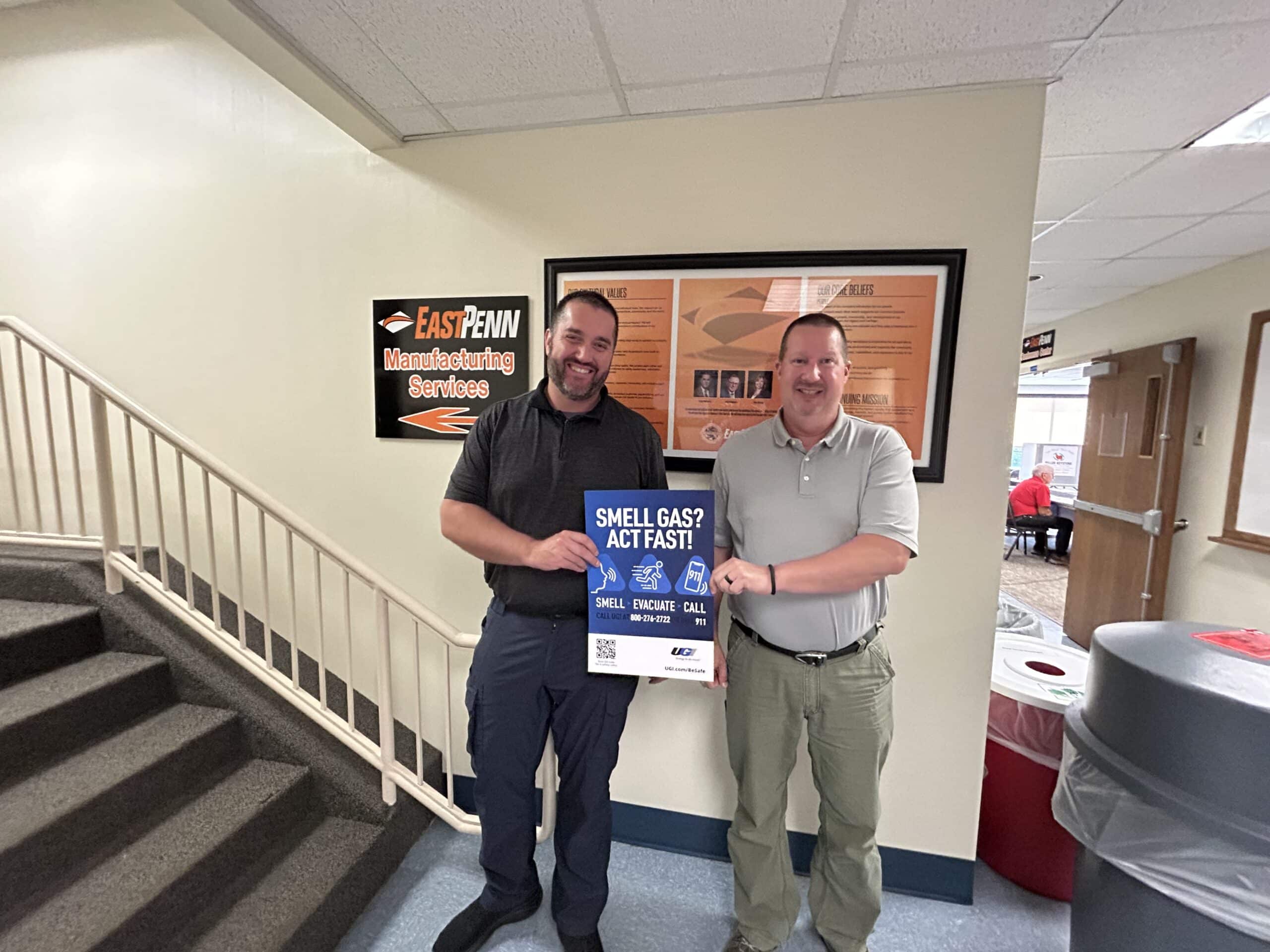 Two men hold poster that says Smell Gas? Act Fast! in front of East Penn Manufacturing Services sign.